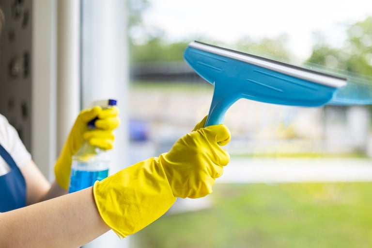 A person is using a blue squeegee to clean a window, holding a spray cleaner in the other hand